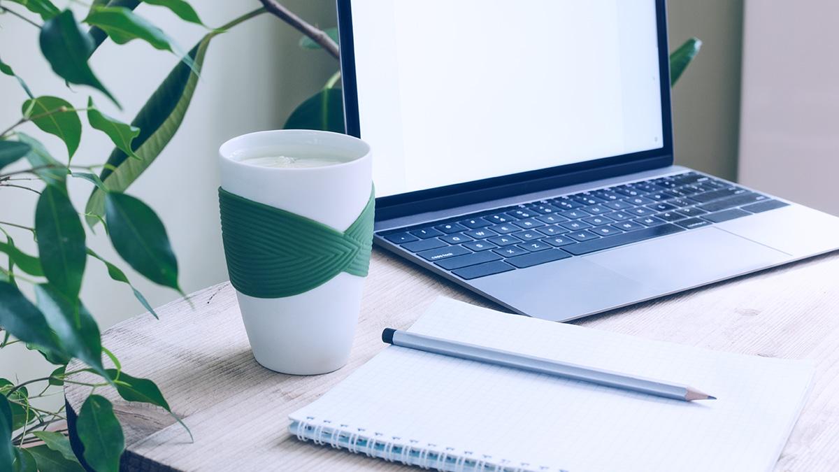 Article Cards Featured Image A modern office workplace surrounded by green plants. A wooden table with a laptop, notebook, pencil and a cup of water and lemon. Desk plants and flowers have many benefits, according to the latest research.