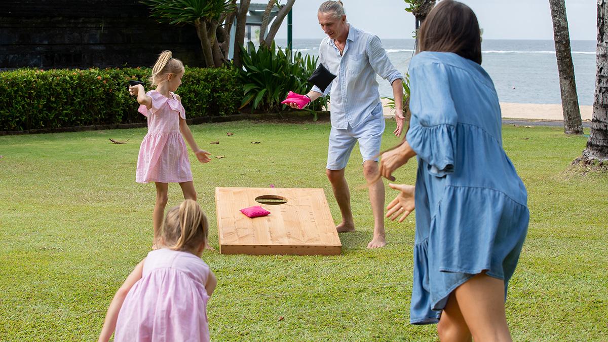 Article Cards Featured Image Happy family playing cornhole game by the sea on sunny summer day. Parents and children playing bean bag toss